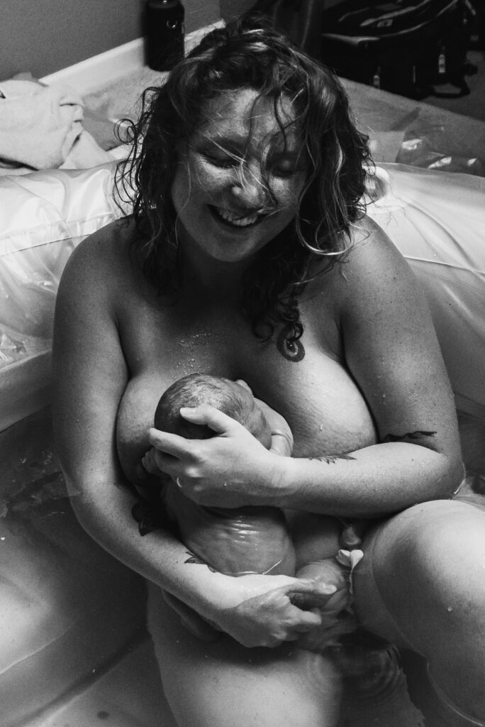 Mom smiling and holding her baby to her chest for the first time in a white birth tub in her home in Johnstown Colorado