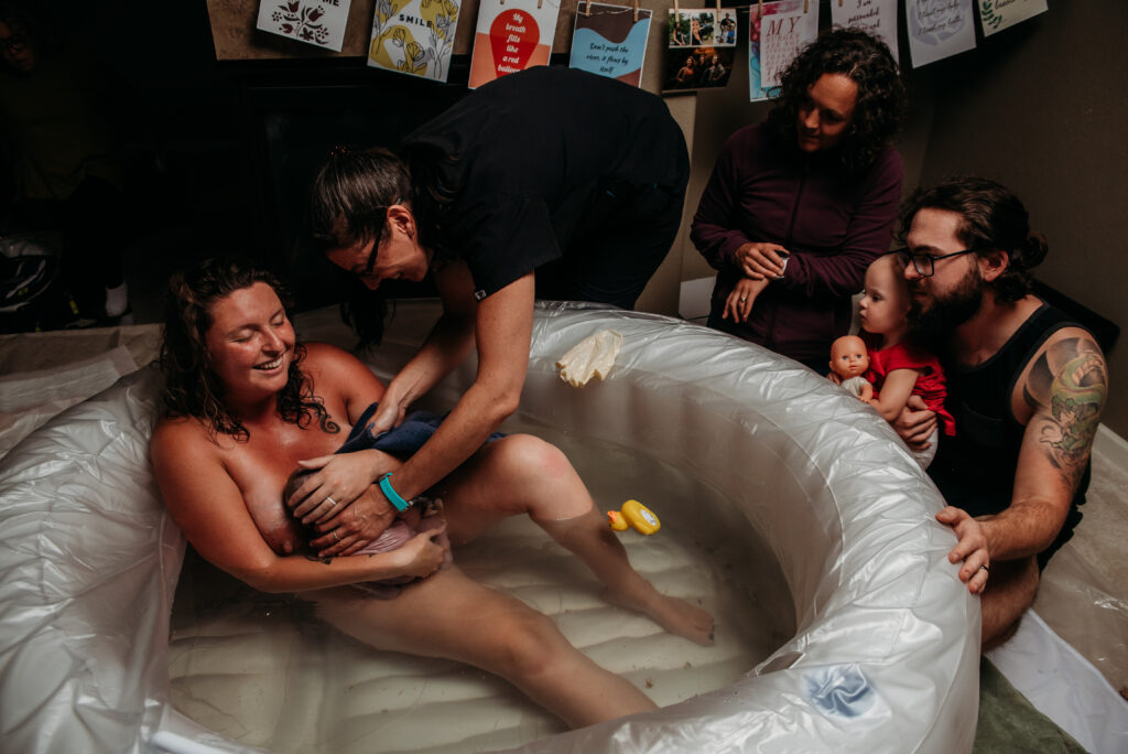 Mom smiling and holding her just born baby in a white birth tub in her home in Johnstown Colorado. Midwife leaning in to assist with baby. Midwife assistant close by. Dad and big sister watching mom and baby.