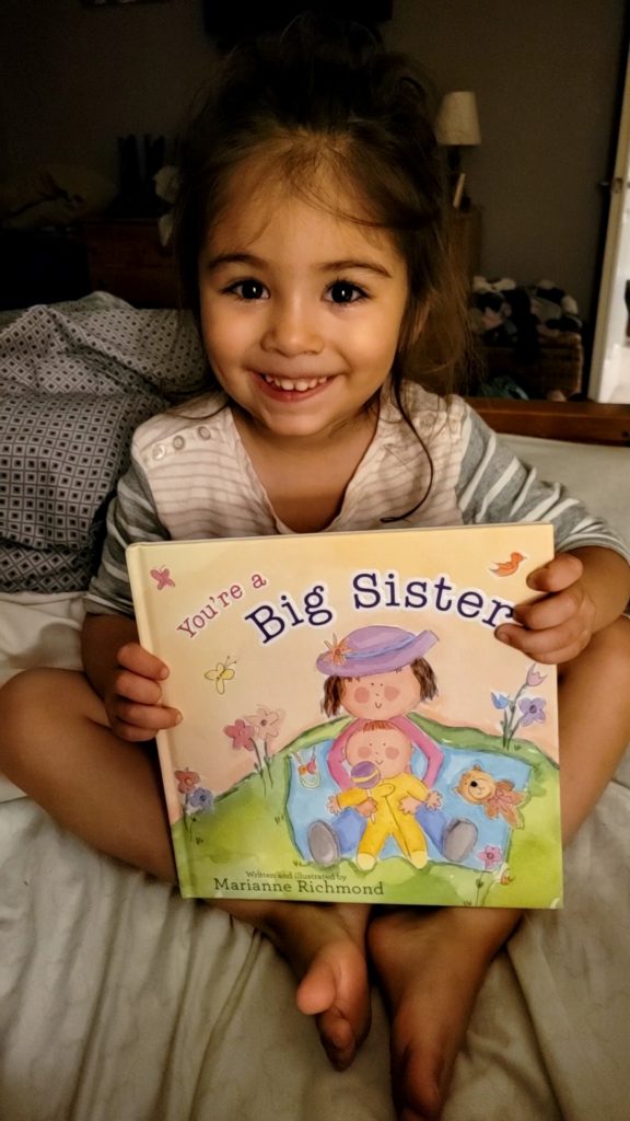 Smiling three year old daughter holding up a book called "You're a big sister"