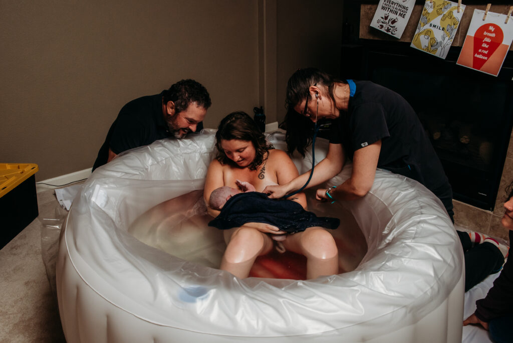Midwife checking just born baby while in the arms of his mom in the white birth pool. Johnstown Colorado home birth photography.