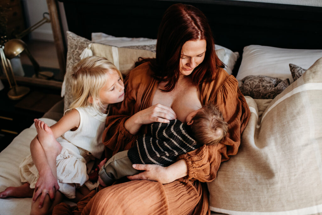 Mom  breastfeeding new baby while big sister looks on and cuddles them on the bed in their home