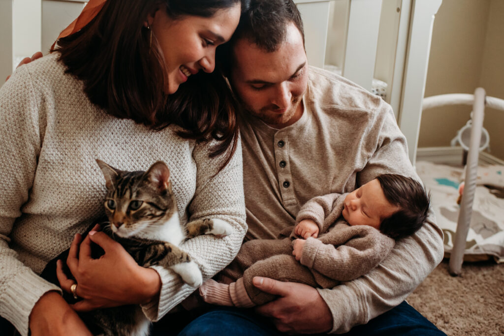 Mom and dad holding new daughter and cat in front of daughter's crib. Lifestyle newborn photo