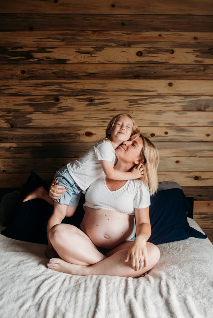 Mom sitting on bed with bare pregnant belly out while her son hugs her. In home maternity photos in Frederick CO