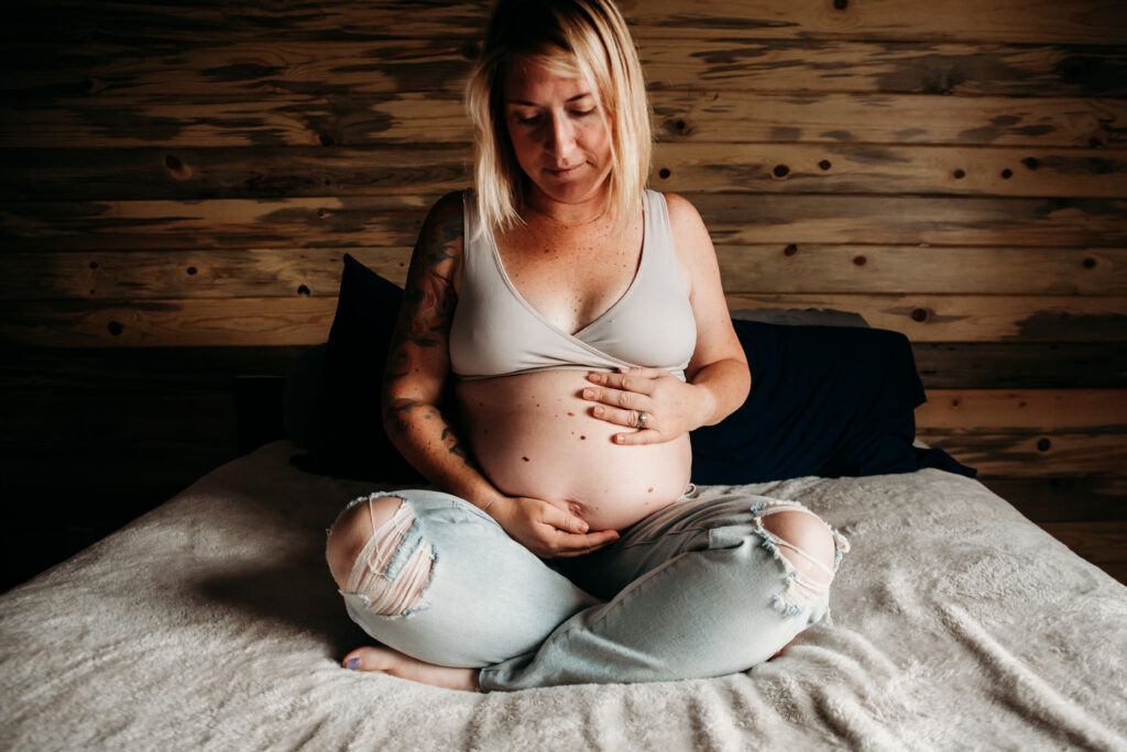 Pregnant mom in ripped jeans and white bra sitting criss cross on the bed during in home maternity photoshoot