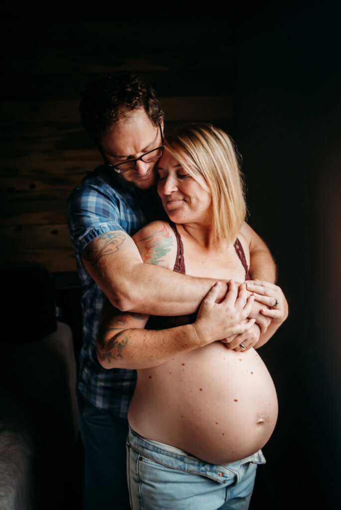 Dad embracing pregnant mom from behind, mom looks back towards dad. Moms bare pregnant belly is out. In home maternity in Frederick CO