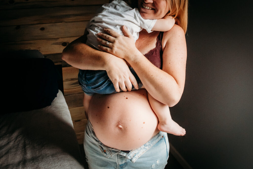 Pregnant bare bellied mom holding her son over her belly during her in home maternity session.