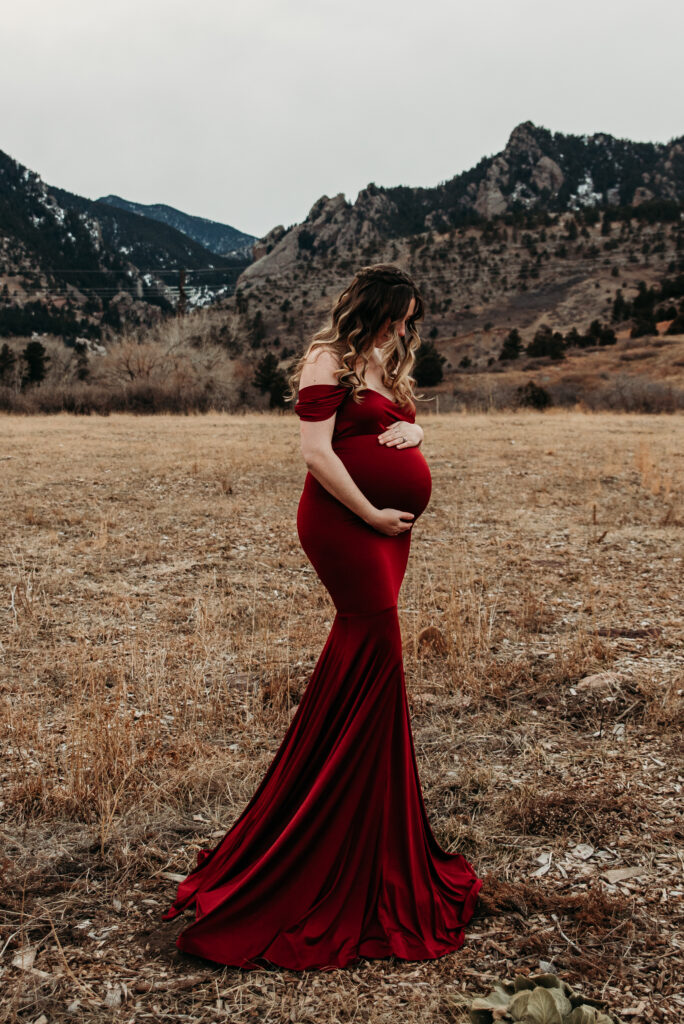 Pregnant mom embracing her belly wearing a long red maternity dress with the Flatirons of Boulder CO in the background