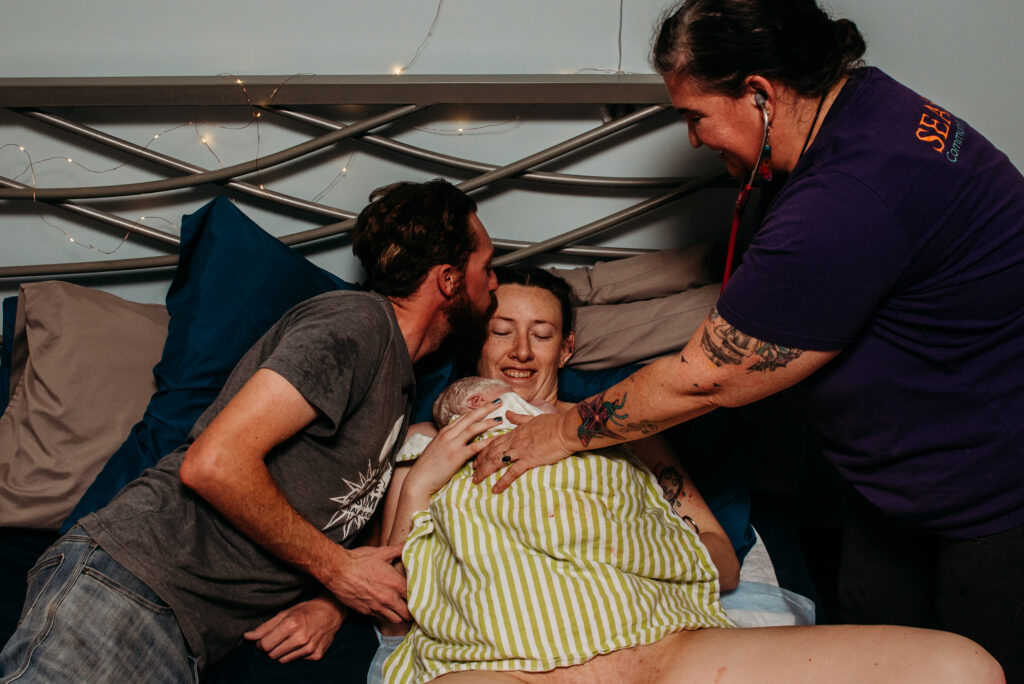 husband kisses wife's forehead while she smiles down at their just born son. Midwife is listening to baby's heart and lungs on the bed at Seasons birth center