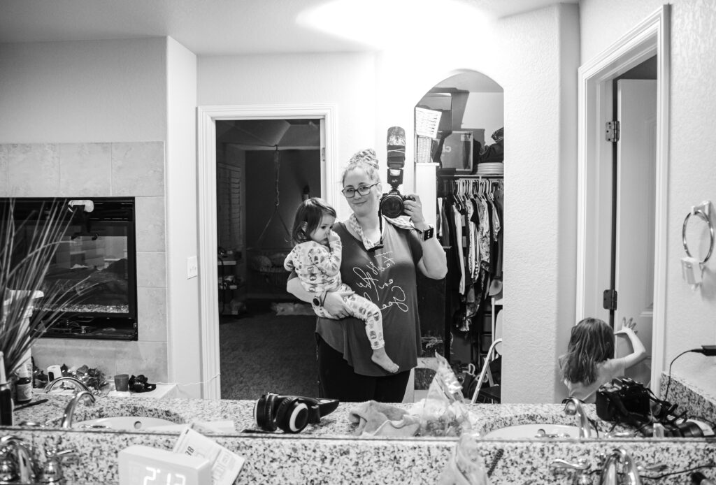 Birth photographer holding client's toddler in one hand and her camera in the other. Mirror selfie.