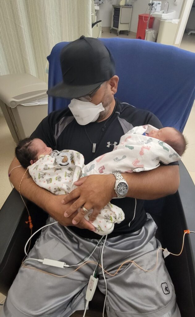 Dad holding both of his twins in the NICU connected to two different machines wearing a mask.