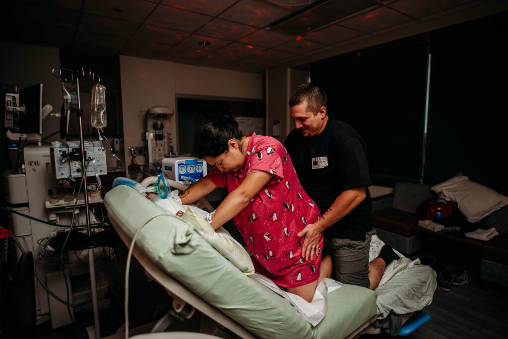 Birth photo of mom laboring on all fours in the hospital bed with husband behind her doing hip squeezes